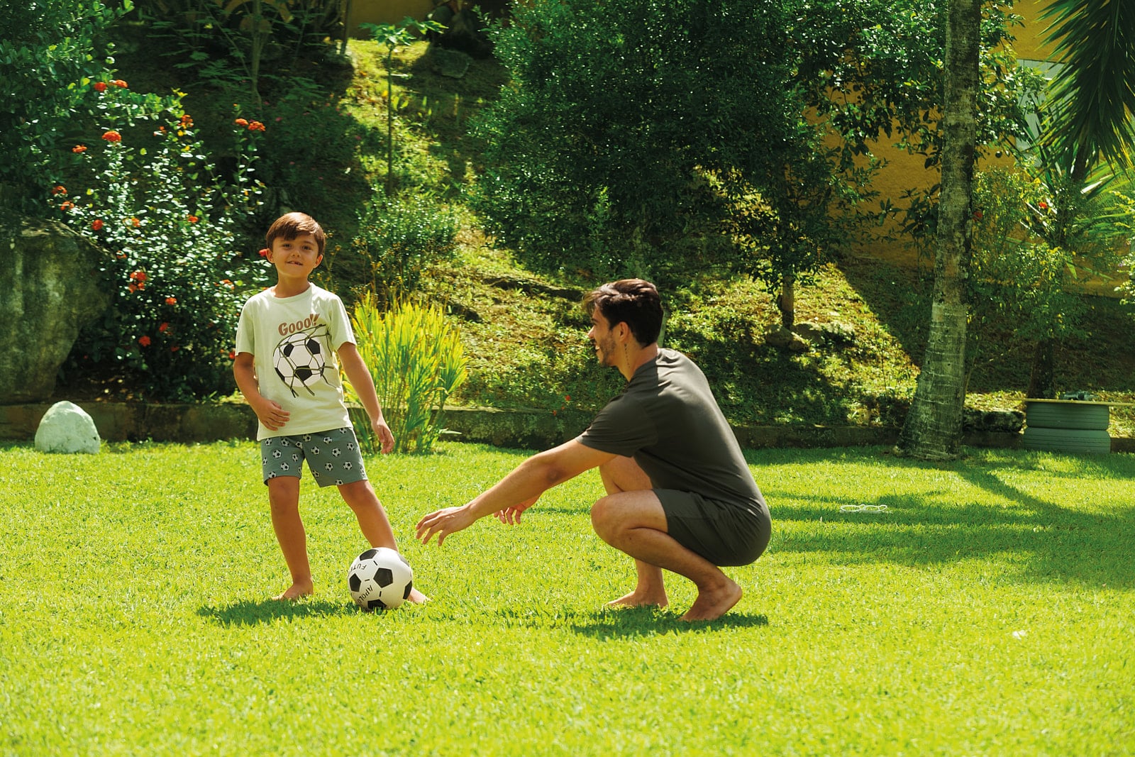 Pai agachado e menino jogando bola no gramado, o menino de camiseta creme com estampa de bola