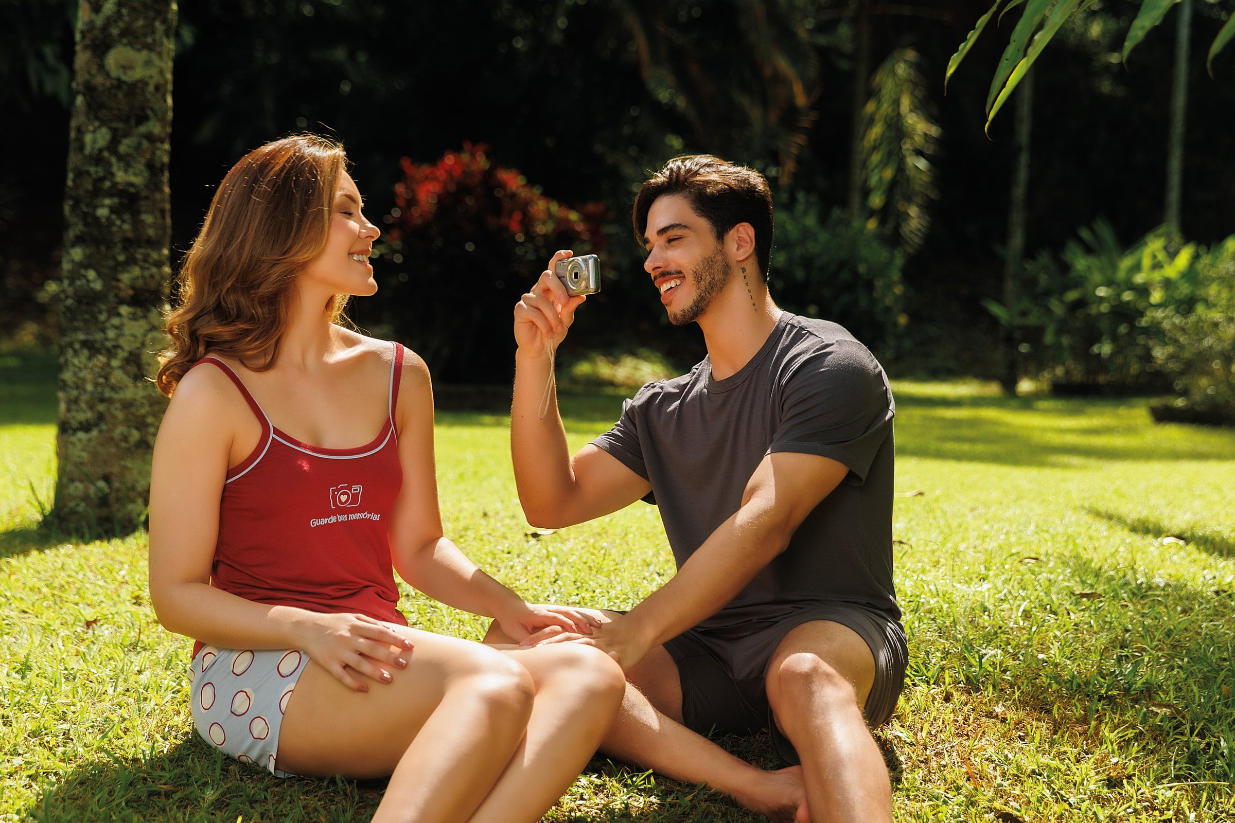 Casal rindo sentado no gramado, ela de regata vermelha e ele de camiseta cinza com uma câmera na mão