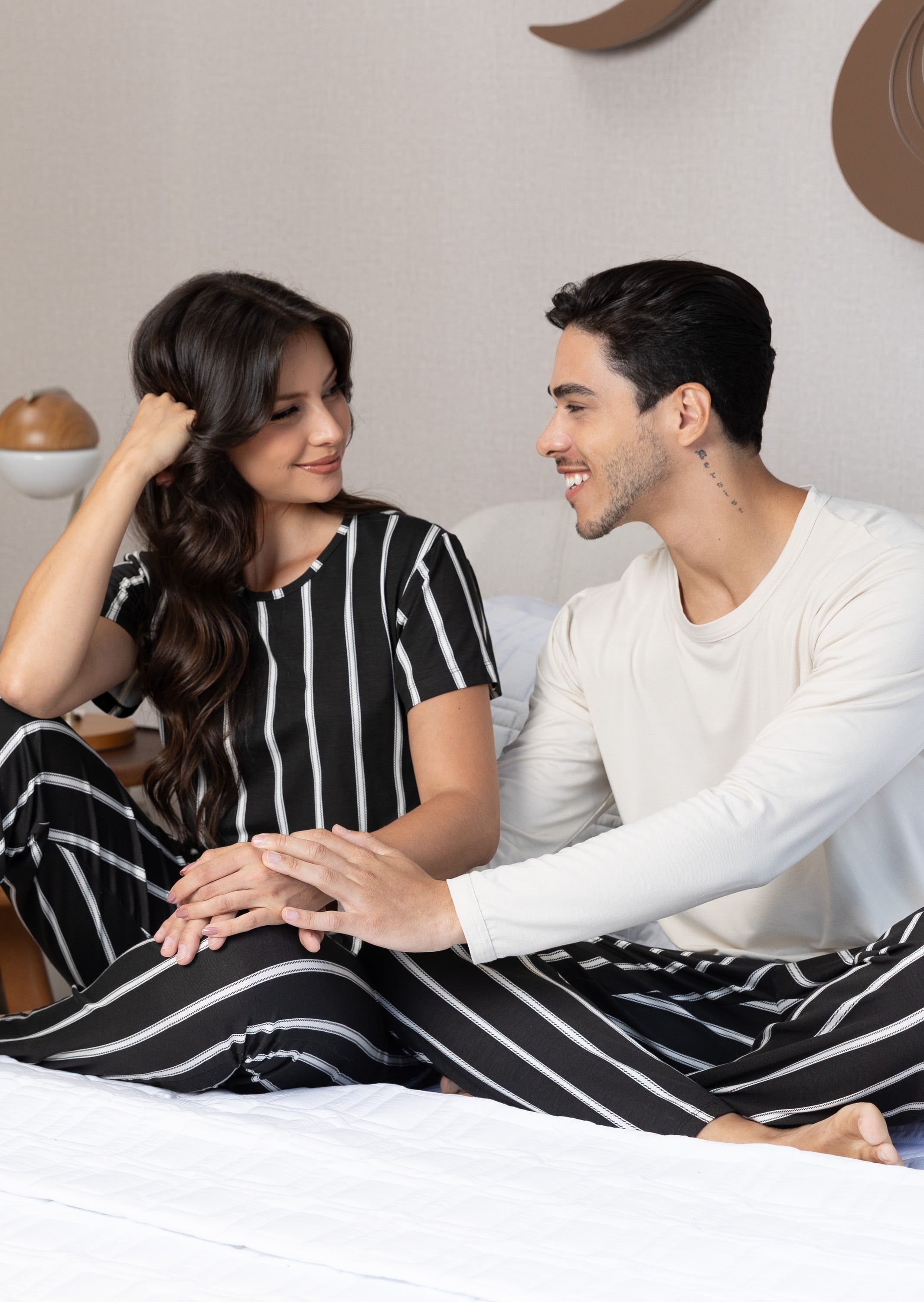 Casal rindo sentado na cama, ela de pijama listrado preto e branco e ele de pijama off-white