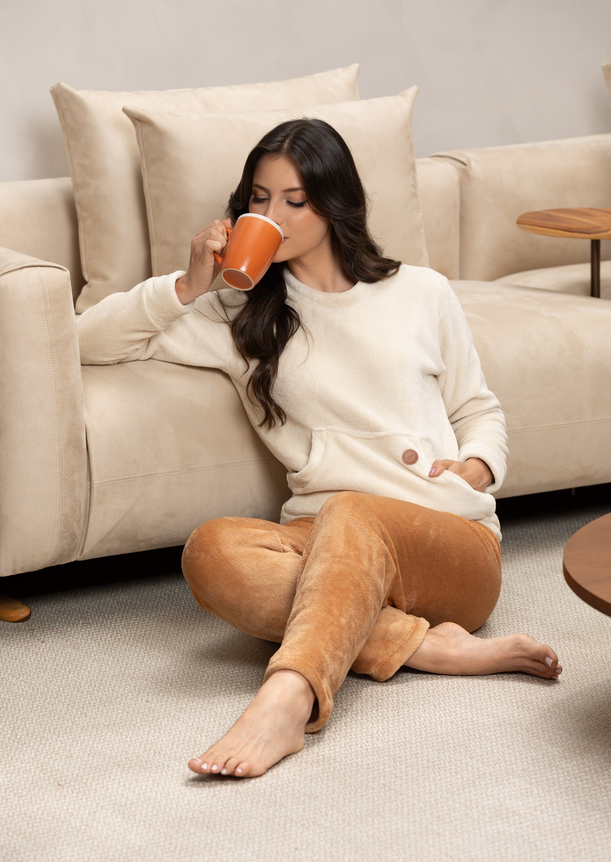 Mulher de blusa creme e calça caramelo tomando café na sala, coleção Sintonia