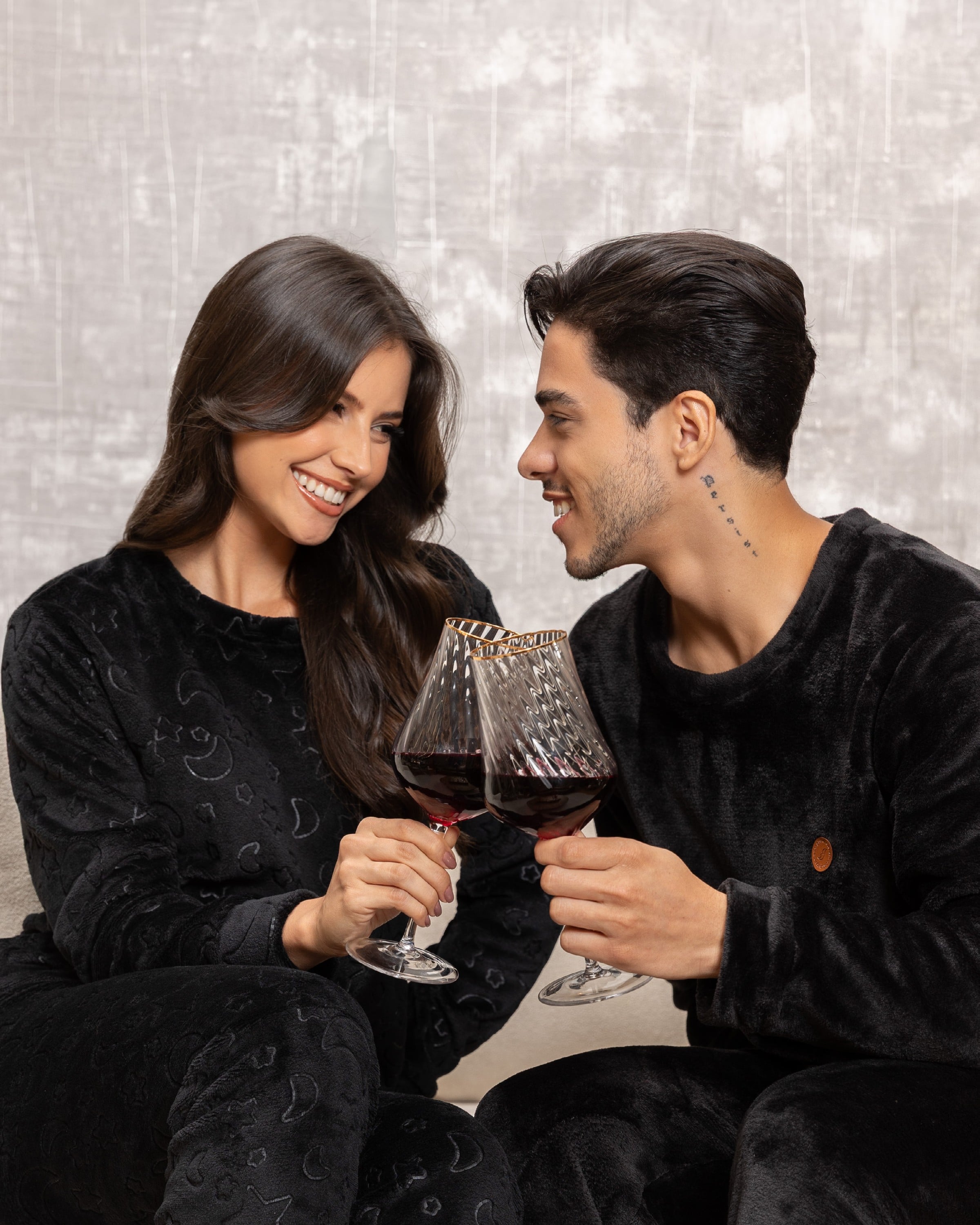 Casal de pijamas pretos de pelúcia brindando com vinho, coleção Sintonia